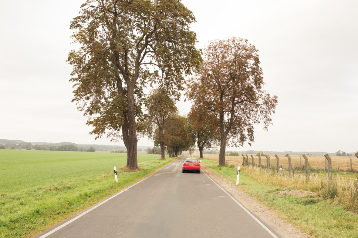 Der rote Audi von Schulz fährt eine Landstraße entlang. Weit und breit nur Wiese und Wald.