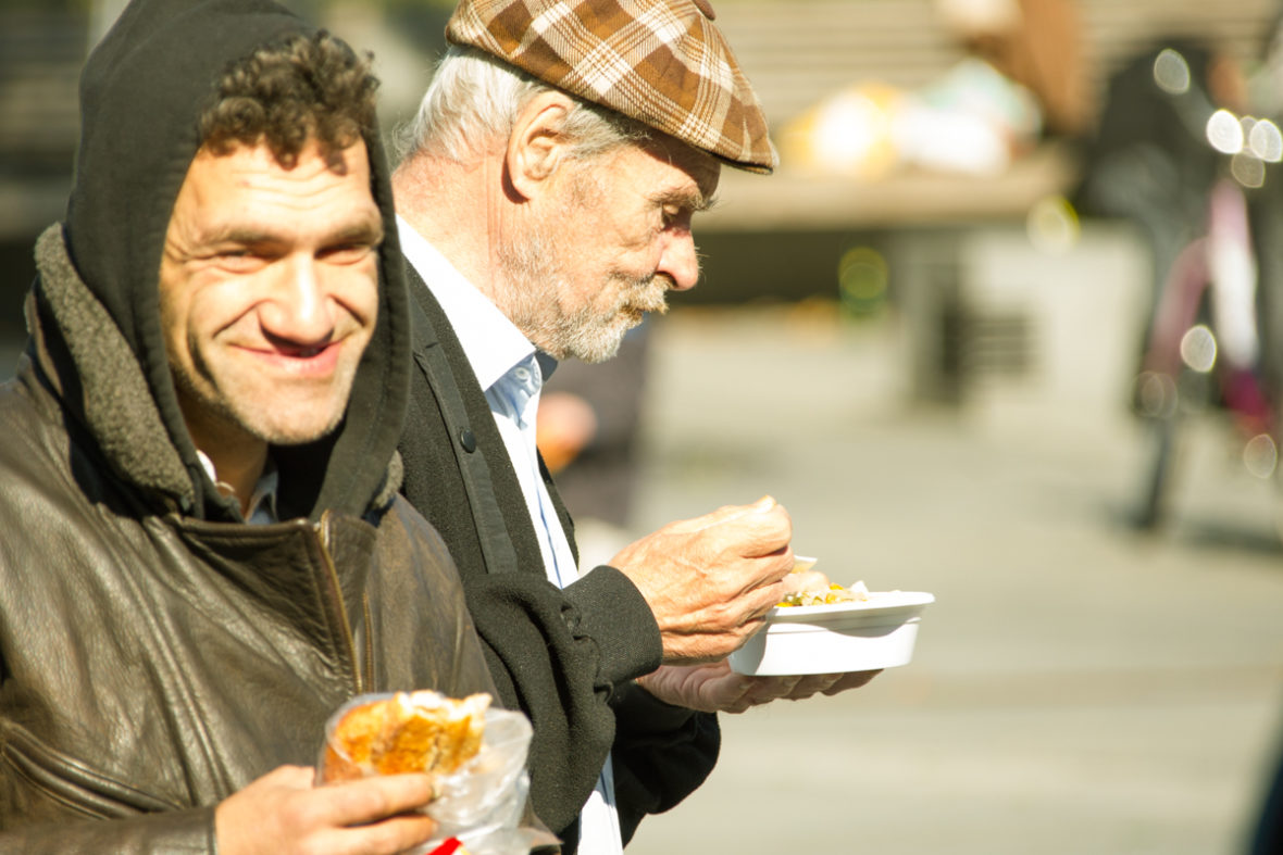 Zwei obdachlose Männer essen gemeinsam, einer lächelt in die Kamera.