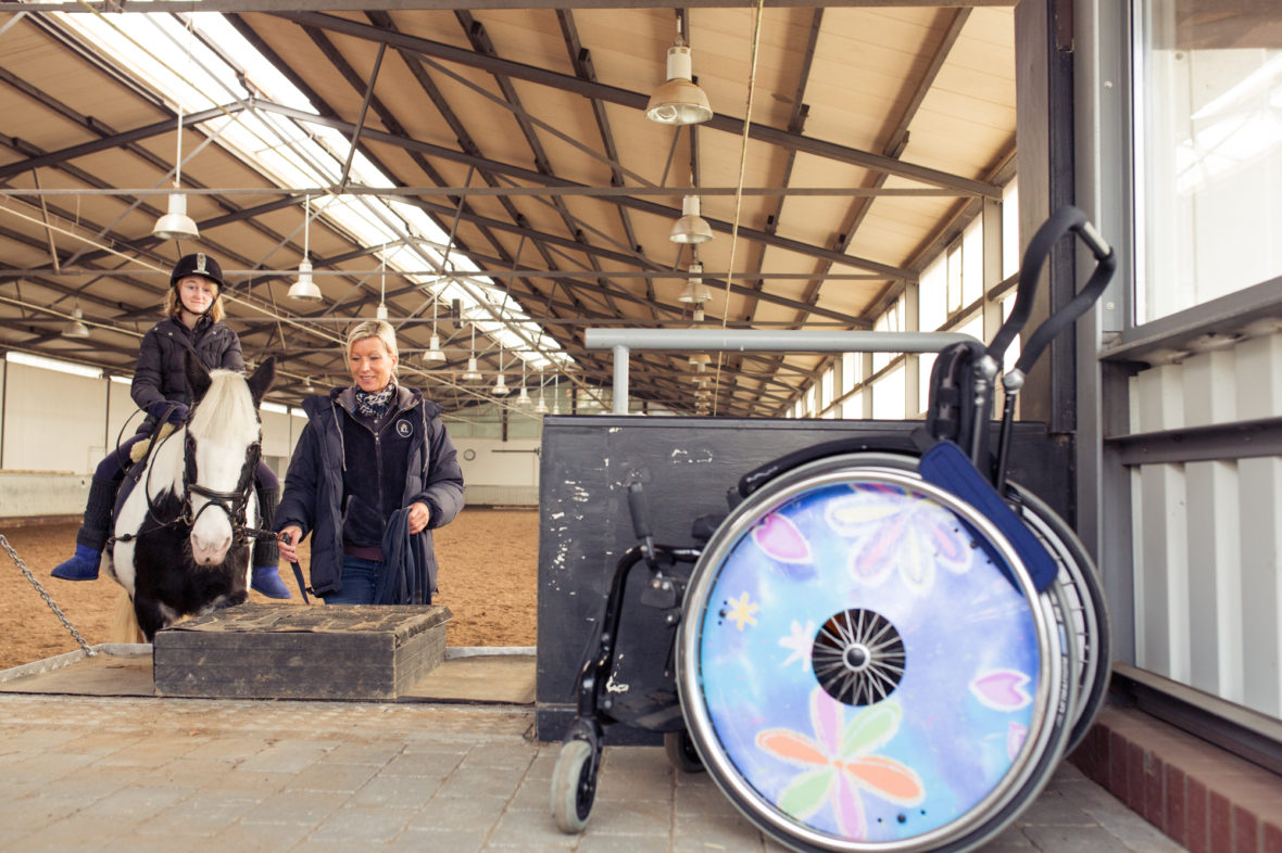 Mächold, Maggy und Alina im Reitstall. Im Vordergrund ist Alina's Rollstuhl zu sehen. Die Radabdeckungen sind bunt.