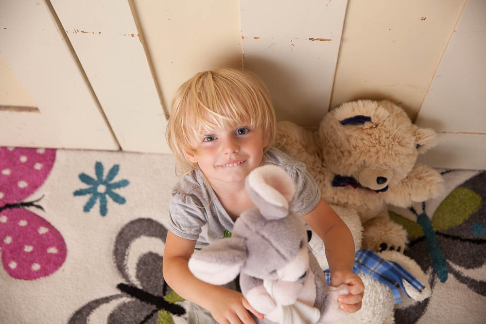 Ein kleiner blonder Junge sitzt auf einem bunten Teppich. Auf seinem Schoß sitzt eine graue Plüschmaus.