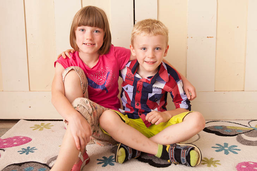 Ein braunhaariges Mädchen mit Pony und ein blonder Junge sitzen nebeneinander auf einem bunten Teppich. Sie legen sich gegenseitig den Arm um die Schulter und lachen in die Kamera.