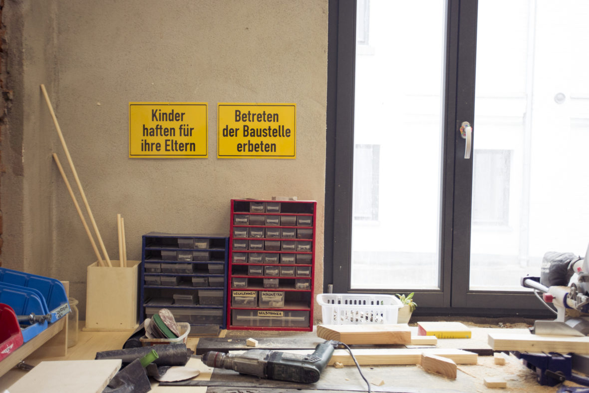 Werkstatttisch: Darüber hängen zwei Schilder. "Kinder haften für Ihre Eltern" und "Betreten der Baustelle erbeten". Auf dem Tisch befinden sich Sortierkästen für Schrauben etc.