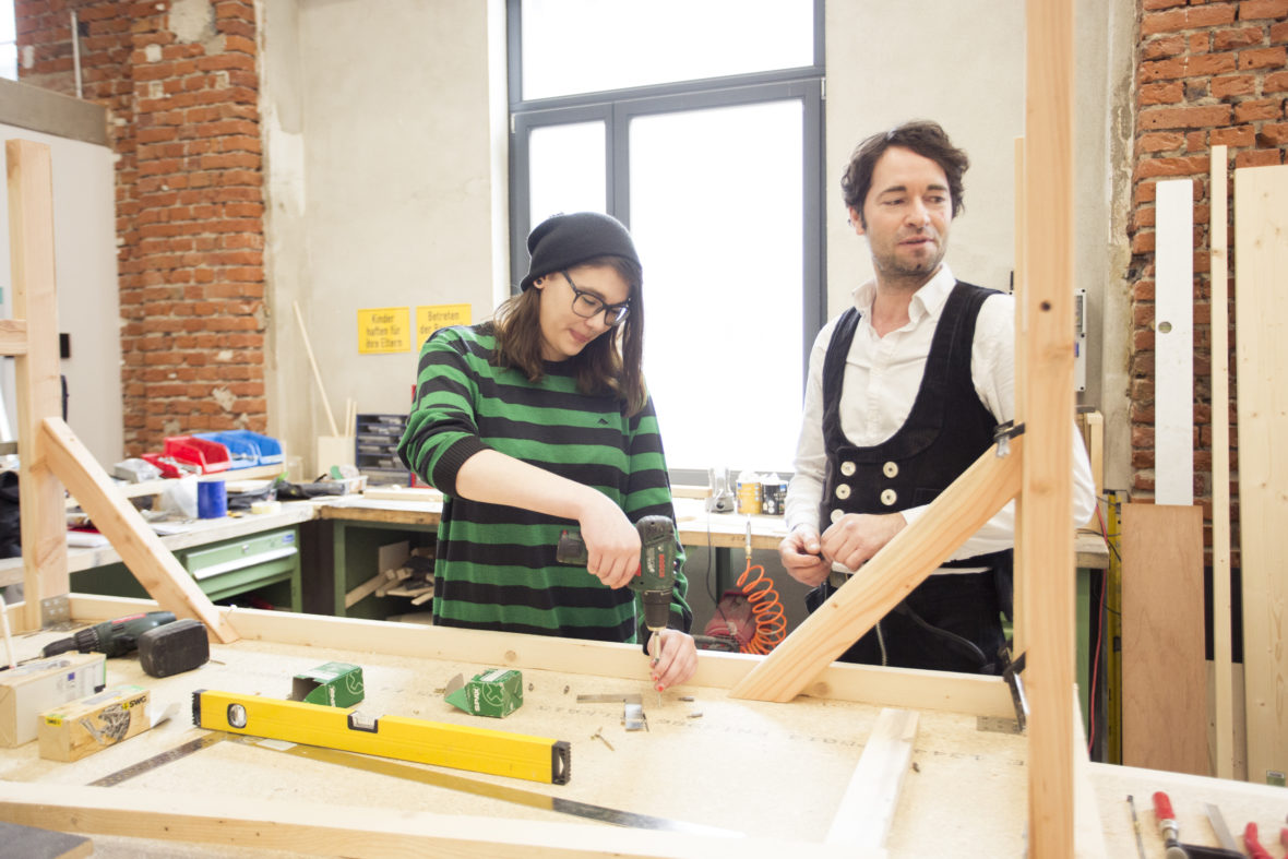 Vanessa arbeitet in der Werkstatt mit einem Bühnenbildner an einem Holzgerüst.