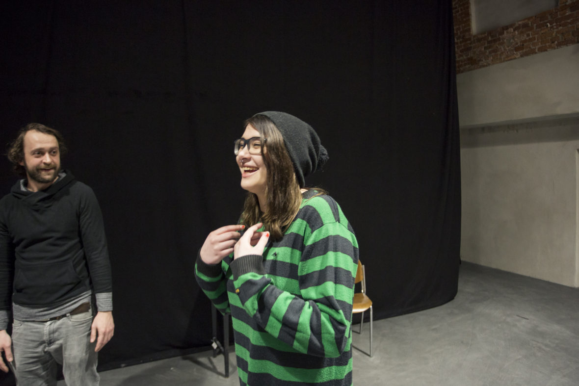 Eine Junge Frau lacht, als sie bei einer Theaterprobe eine Mimik-Übung macht. Ihr Betreuer sieht zu