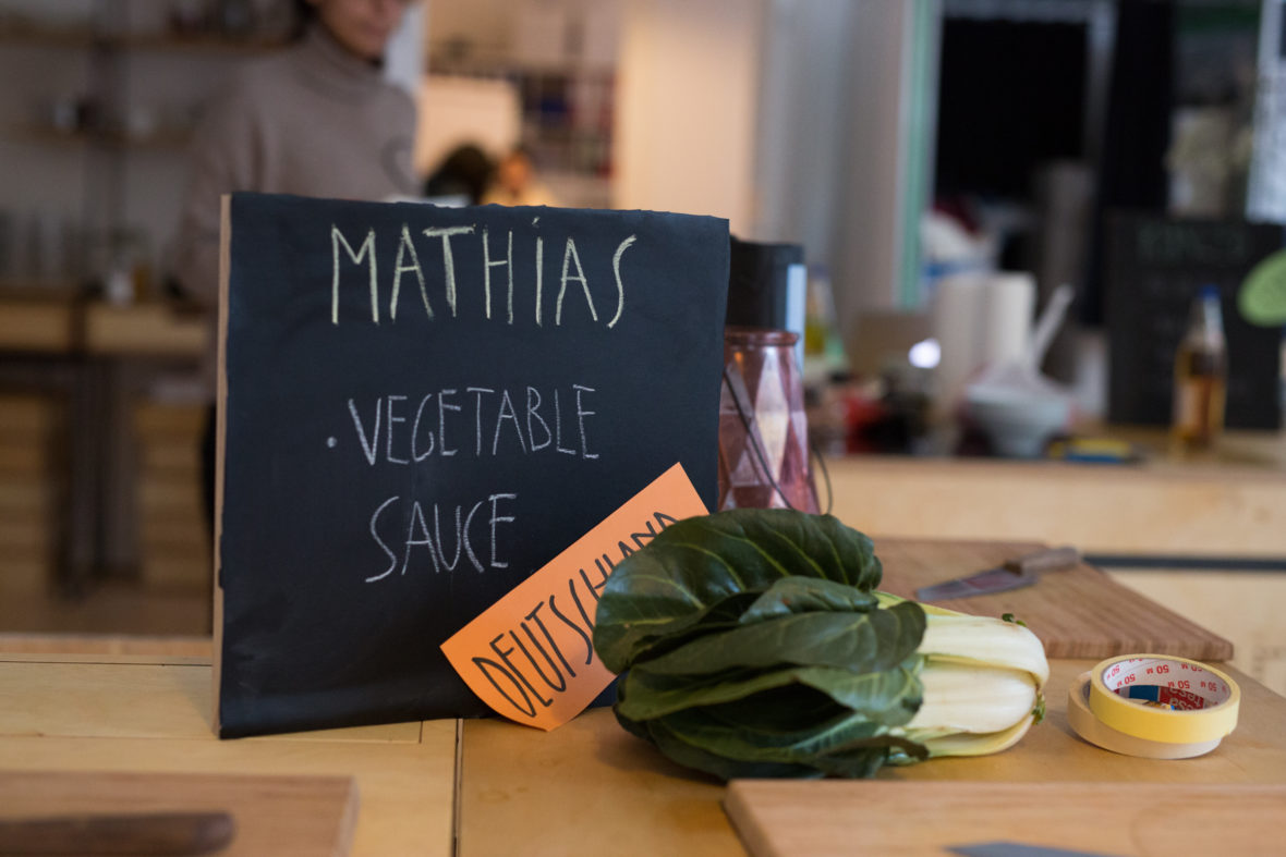 Eine Tafel, auf der der Name Mathias steht und darunter auf Englisch: Gemüsesoße. Halb davor liegt ein Kopf Salat.