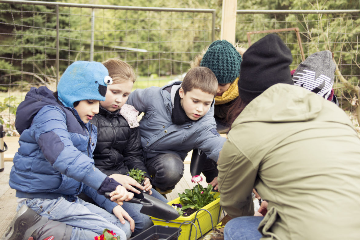 Drei Kinder pflanzen Blumen in einen gelben Blumenkasten.