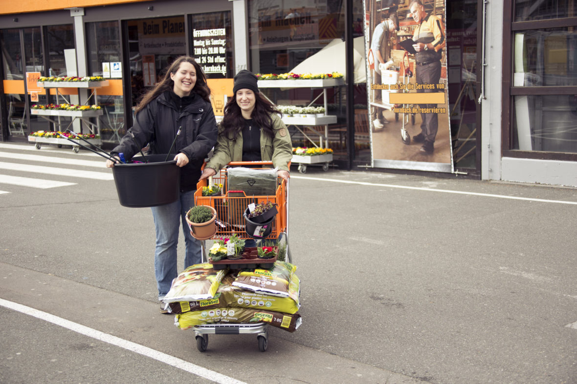 Katharina Kildau und Lena Schmitt schieben einen Einkaufswagen voller Blumen und Blumendere über einen Parkplatz.