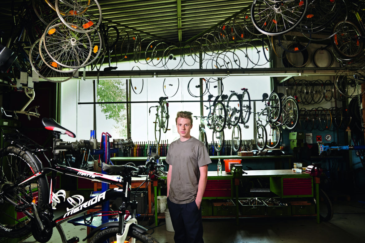 Ein junger Mann steht, die Hände in den Taschen, in einer Werkstatt, voller Fahrrad-Räder.
