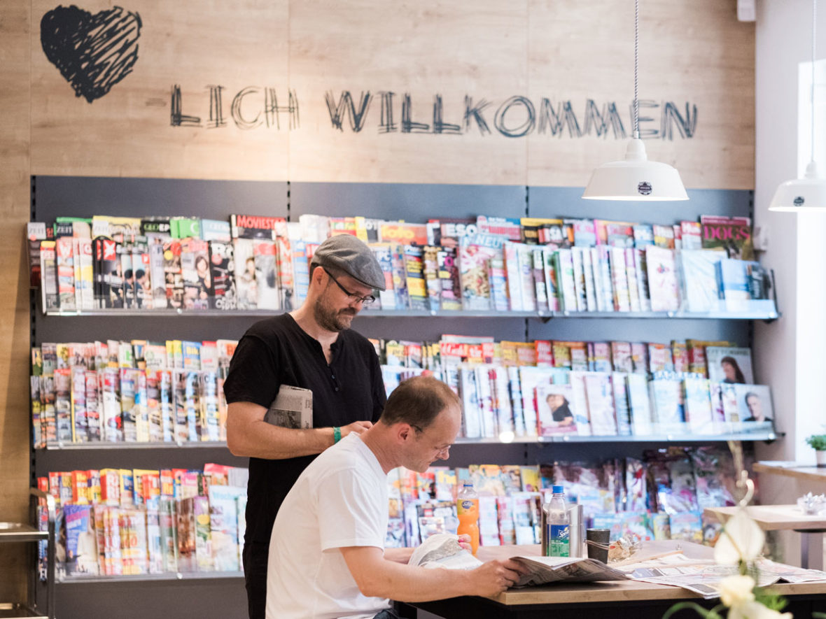 Ein Mann mit einer Baskenmütze steht vor einem Zeitschriftenregal und liest, ein anderer sitzt an einem Tisch und schaut in die Zeitung.