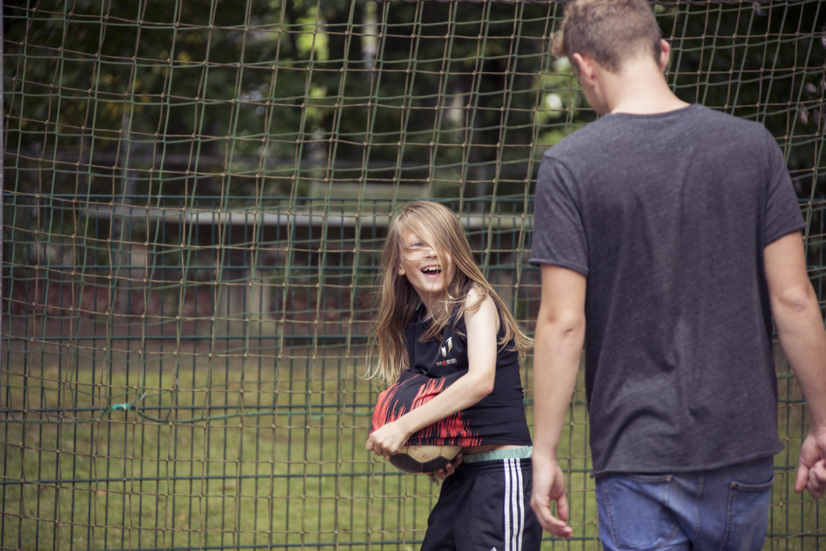 Ein Junge mit langen Haaren versteckt einen Fußball unter seinem T-Shirt und lacht. Ein Erwachsener will ihm neckisch den Ball abnehmen.
