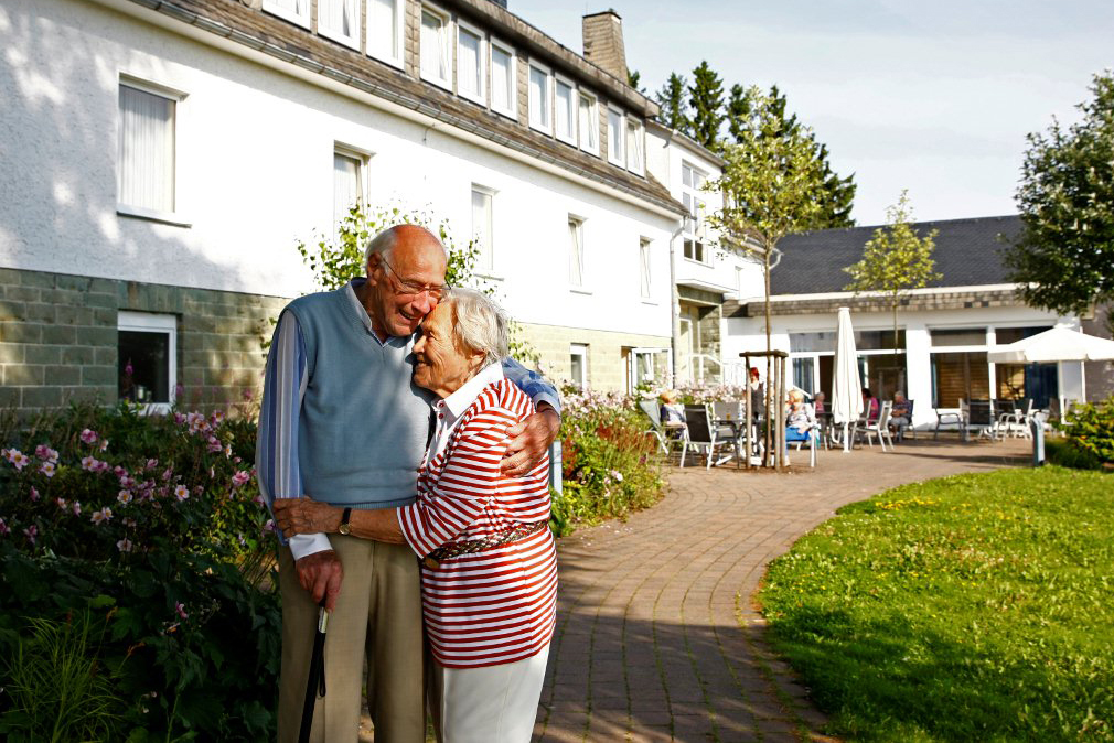 Ein alter Mann und eine alte Frau umarmen sich innig im Hof eines Pflegeheims.