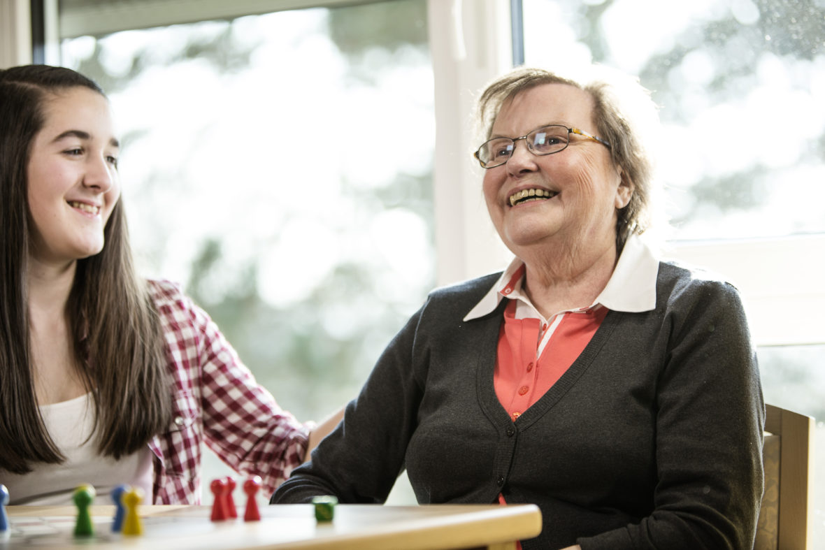 Eine junge Frau streicht einer älteren Dame über den Arm, beide lächeln.