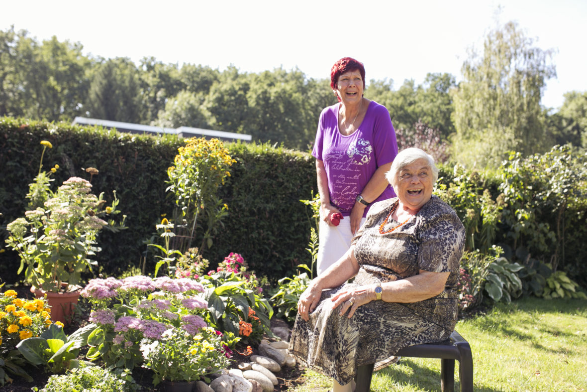 Zwei Frauen, eine ältere auf dem Stuhl und eine mit roten Haaren sind im Garten und lachen von Herzen.