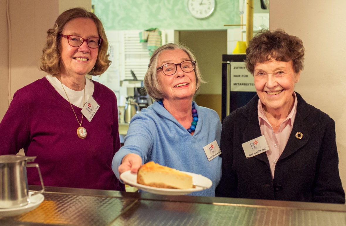 Drei ältere Damen stehen an einem Tresen, eine von ihnen hält einen Kuchen auf einem Teller. Sie lächeln.