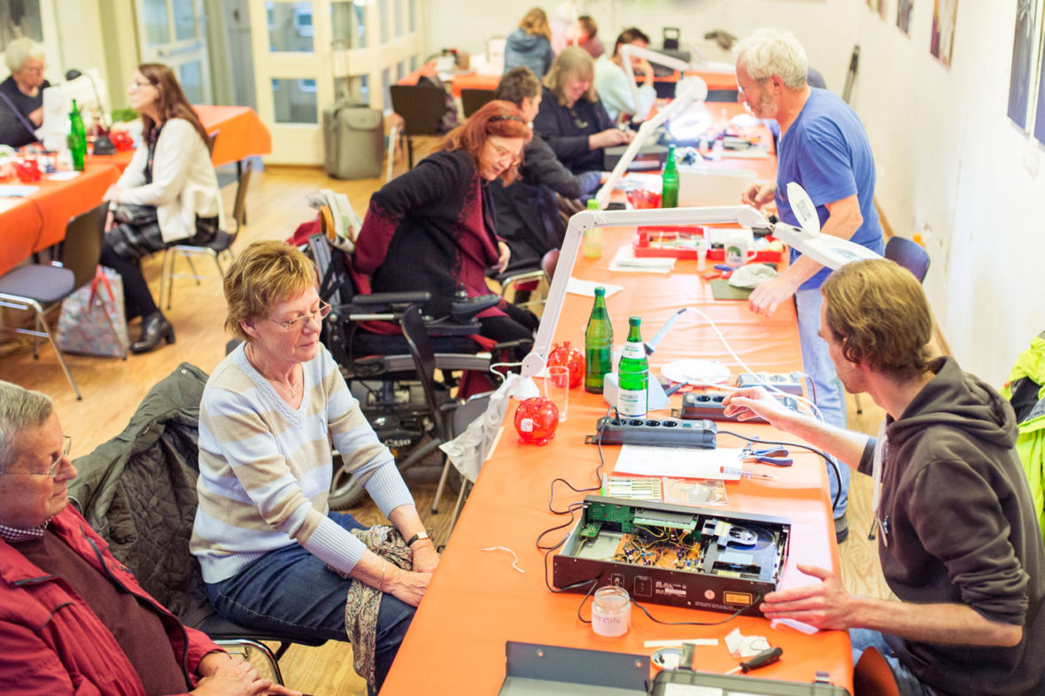 An langen Tischen sitzen ältere und jüngere Leute. Die Helfer sitzen ihnen gegenüber und arbeiten an den defekten Geräten, die die Besucher mitgebracht haben.