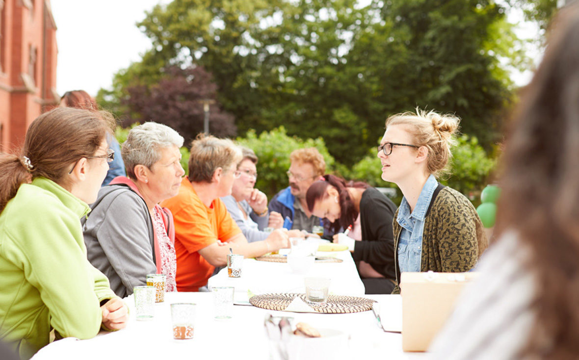 Alle sitzen gemeinsam an einer langen Tee-Tafel und unterhalten sich.