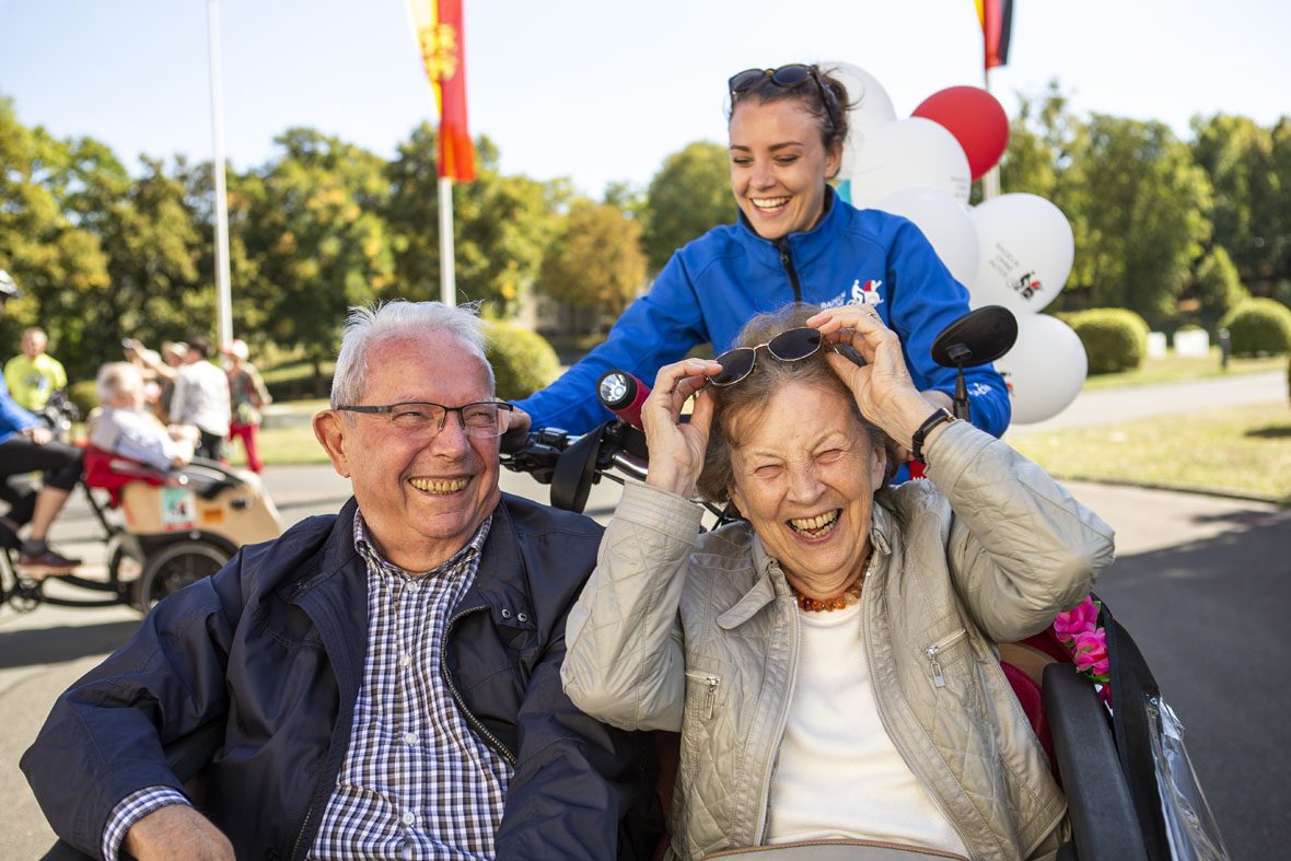 Der ältere Herr und die ältere Dame in der Rikscha lachen, die Dame greift nach ihrer Sonnenbrille.
