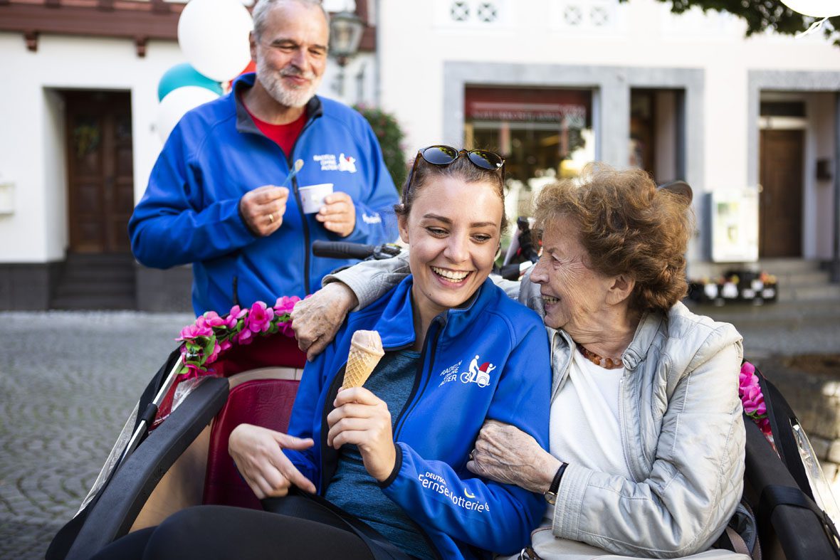 Caroline Kuhl sitzt mit einer Seniorin in der Rikscha, die Seniorin umarmt die Studentin und schaut sie lachend an.