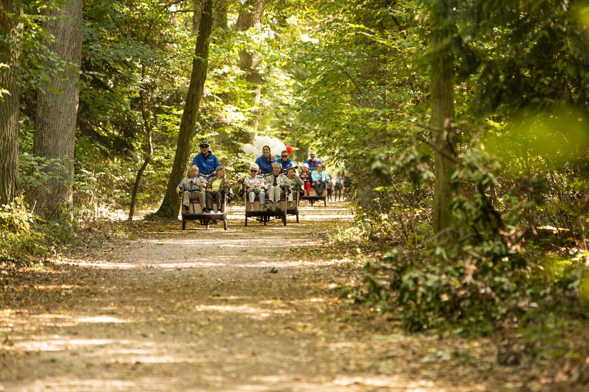 Auf einem Waldweg kommen in der Ferne vier Fahrradrikschas, geschmückt mit bunten Luftballons, mit je einem Fahrerer/einer Fahrerein und zwei Senioren/Seniorinnen angefahren.