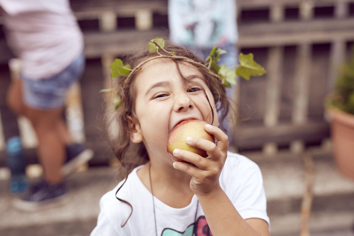 Ein Mädchen mit einem Efeukranz auf dem Kopf beißt in einen Apfel.