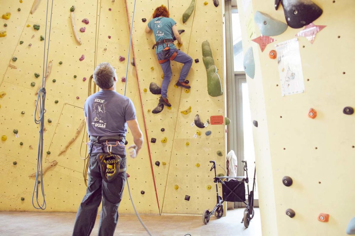 Sanna klettert an der Wand, rechts unter ihr auf dem Boden steht ihr Rollator. Ein Helfer sichert sie.
