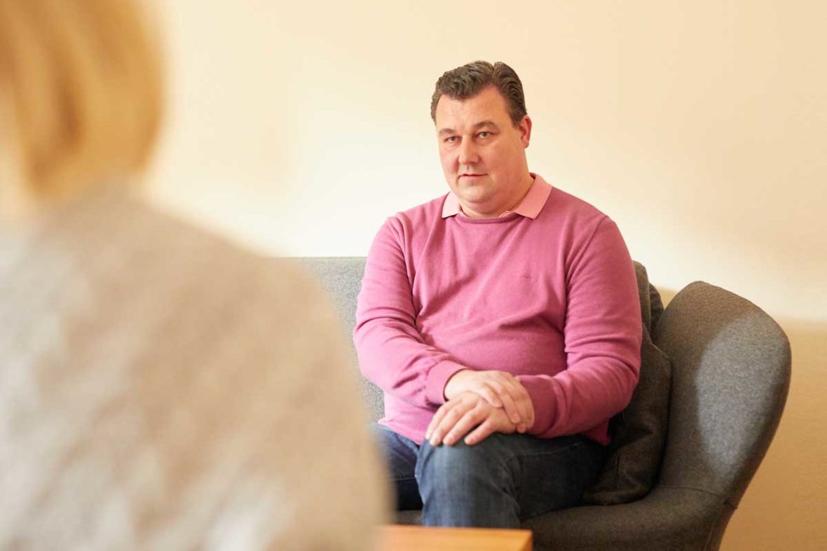 Michael Rieke sitzt auf einem grauen Sofa im Lotsenhaus. Er hat die Hände gefaltet auf sein linkes Bein gelegt.