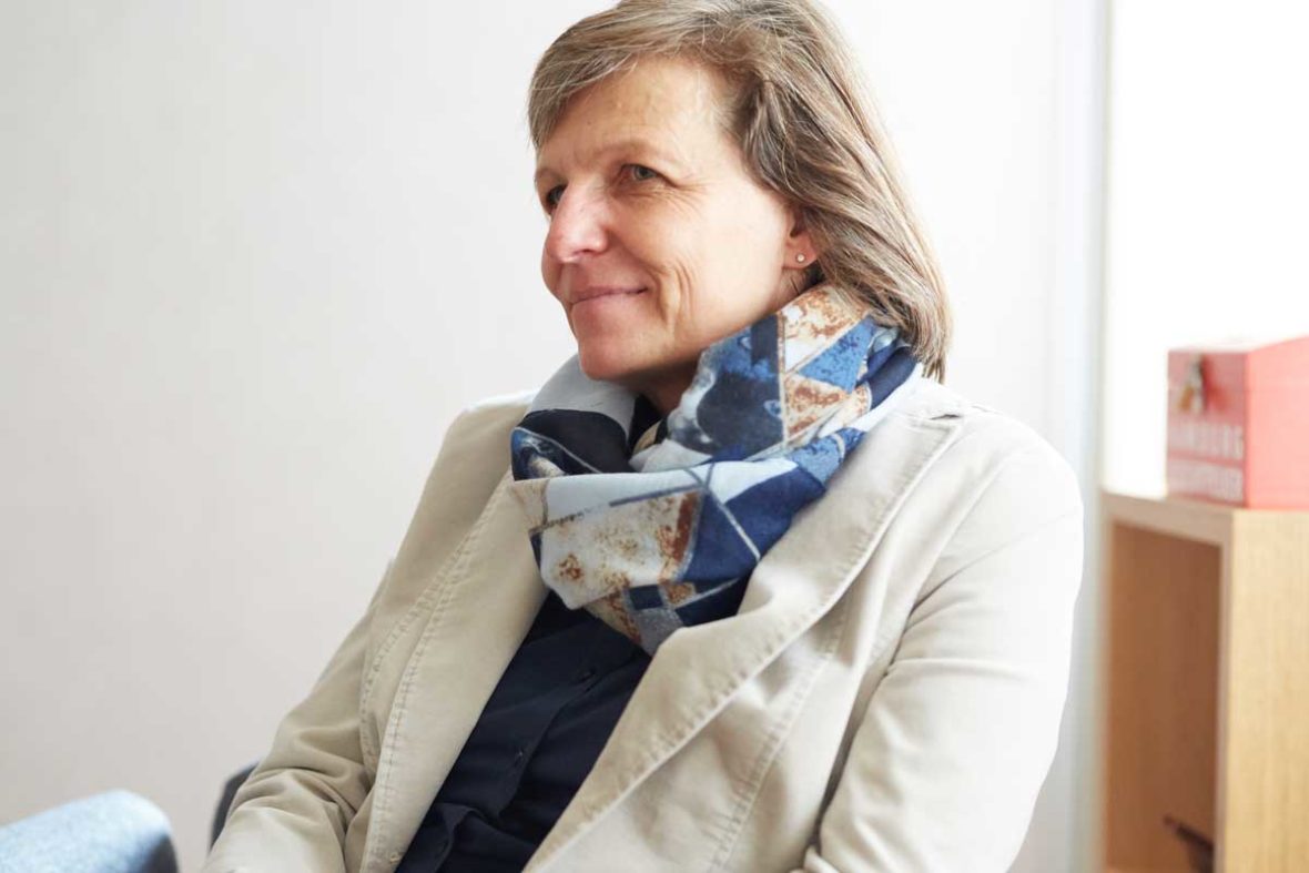 Bettina Kriegel sitzt auf einem Sessel im Lotsenhaus und schaut freundlich an der Kamera vorbei, in die Richtung, wo Michael Rieke sitzt. Sie trägt einen weißen Blazer und einen blaugemusterten Schal.