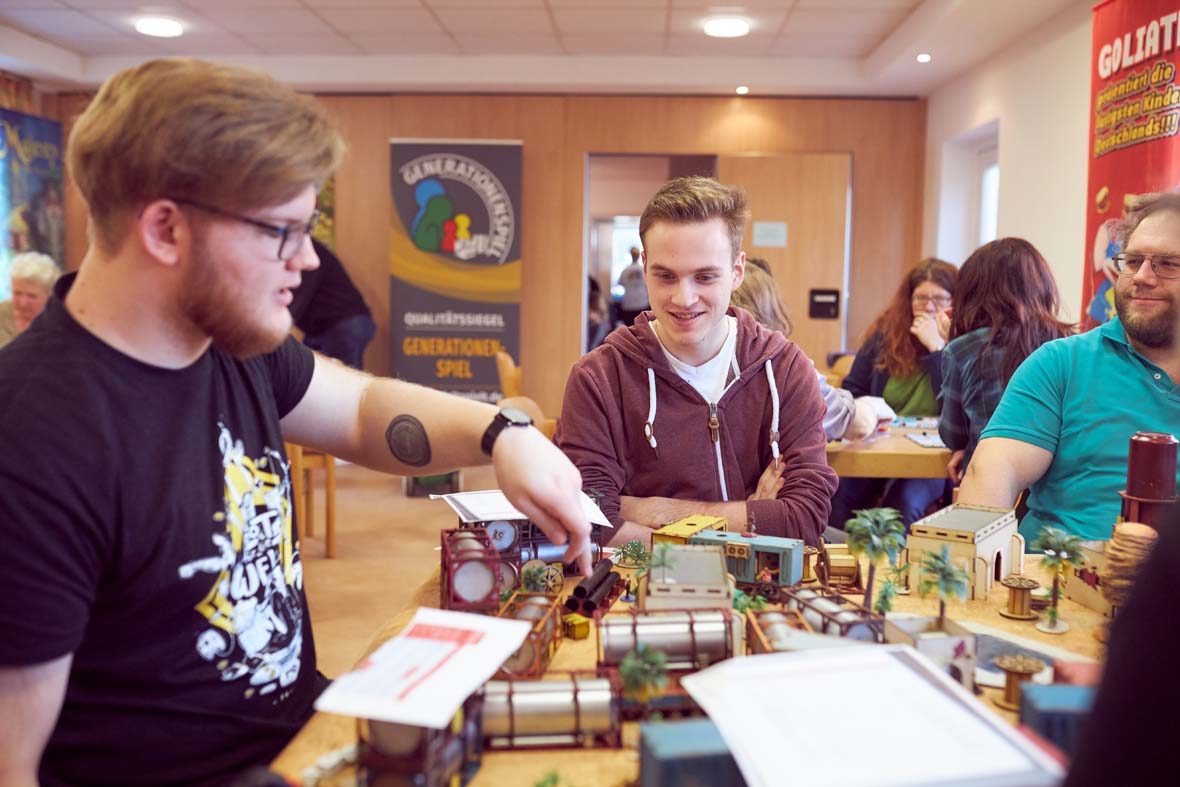 Eine gruppe junger Männer spielt ein Rollenspiel, auf dem Tisch steht eine richtige kleine Landschaft mit Plastikpalmen, Gebäuden und Containern.