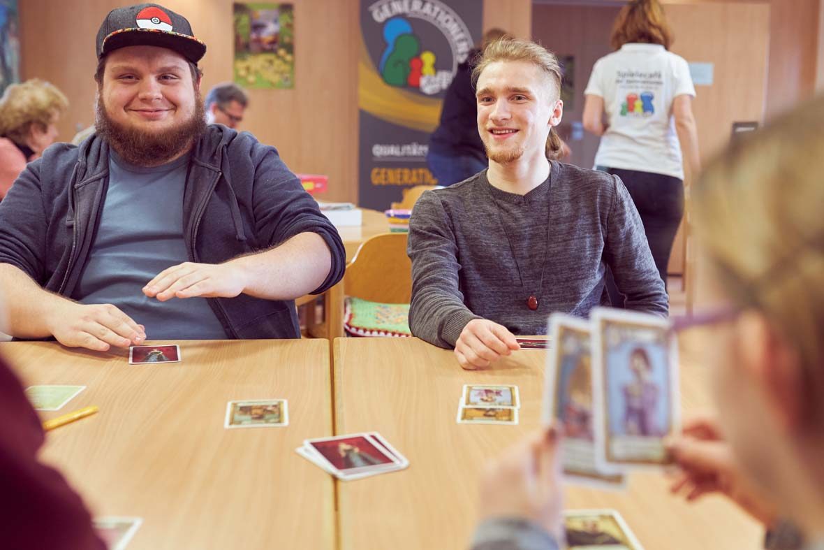 Zwei junge Männer sitzen am Tisch und spielen Karten, ihnen gegenüber sitzt ein junges Mädchen, dessen Kopf und Spielkarten verschwommen im Bild zu sehen sind.