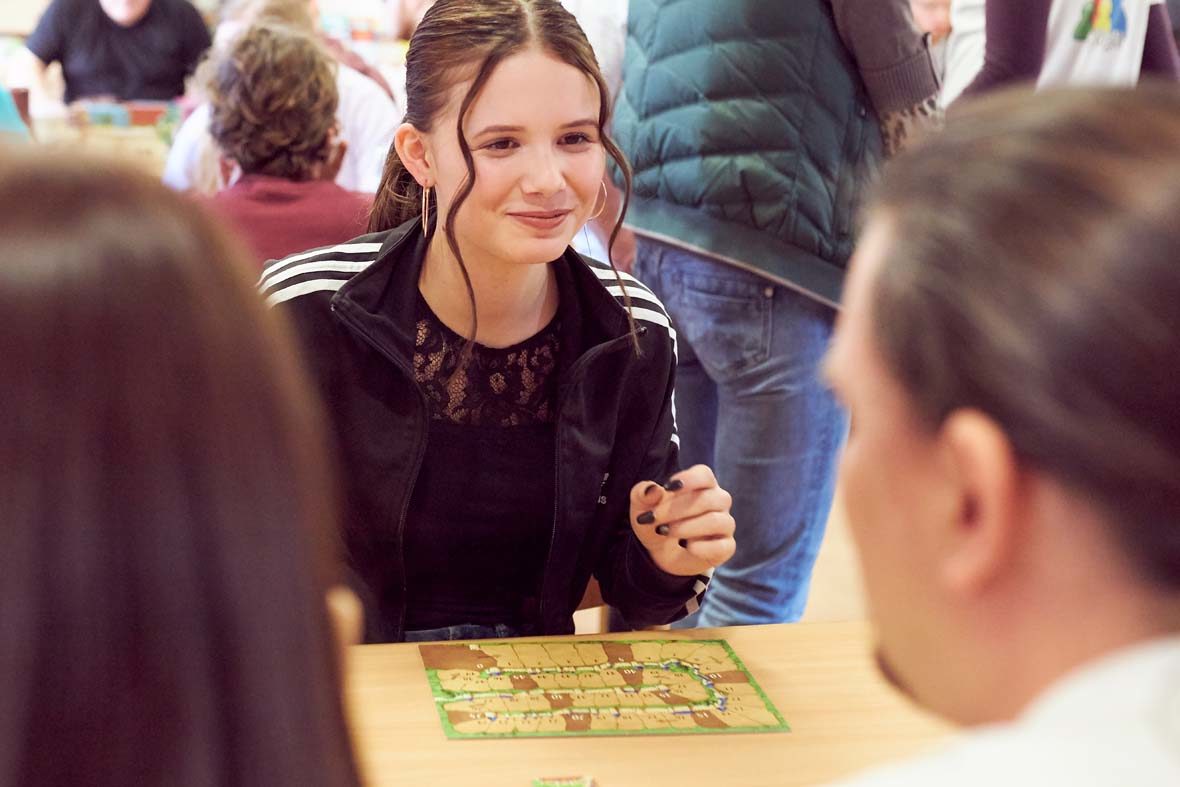 Eine junge Frau mit braunen Haaren und Pferdeschwanz sitzt vor einem kleinen Spielbrett. Sie schaut ihr Gegenüber lächelnd an.