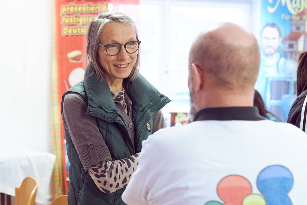 Eine Frau mit kinnlangen grauen Haaren und Brille unterhält sich mit einem Ehrenamtlichen des Vereins, der mit dem Rücken zur Kamera steht.