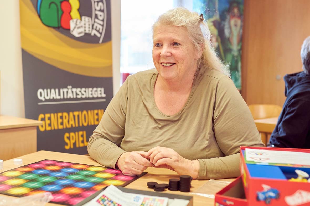 Eine ältere Frau mit blonden Haaren sitzt vor einem bunten Spielbrett. Sie schaut ihren Gegenüber an, den man im Bild aber nicht sieht.