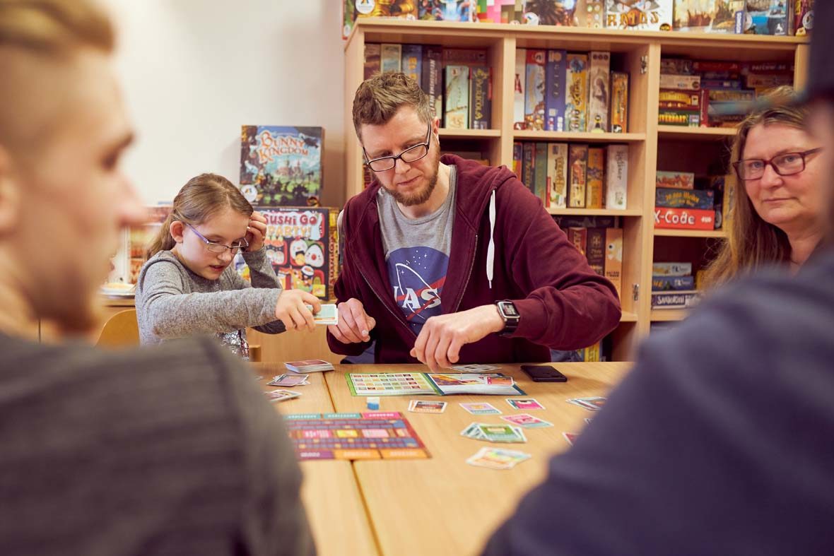 Ein junges Mädchen und ein junger Mann spielen ein Brettspiel, hinter ihnen ist ein Regal voller Spiele zu sehen.