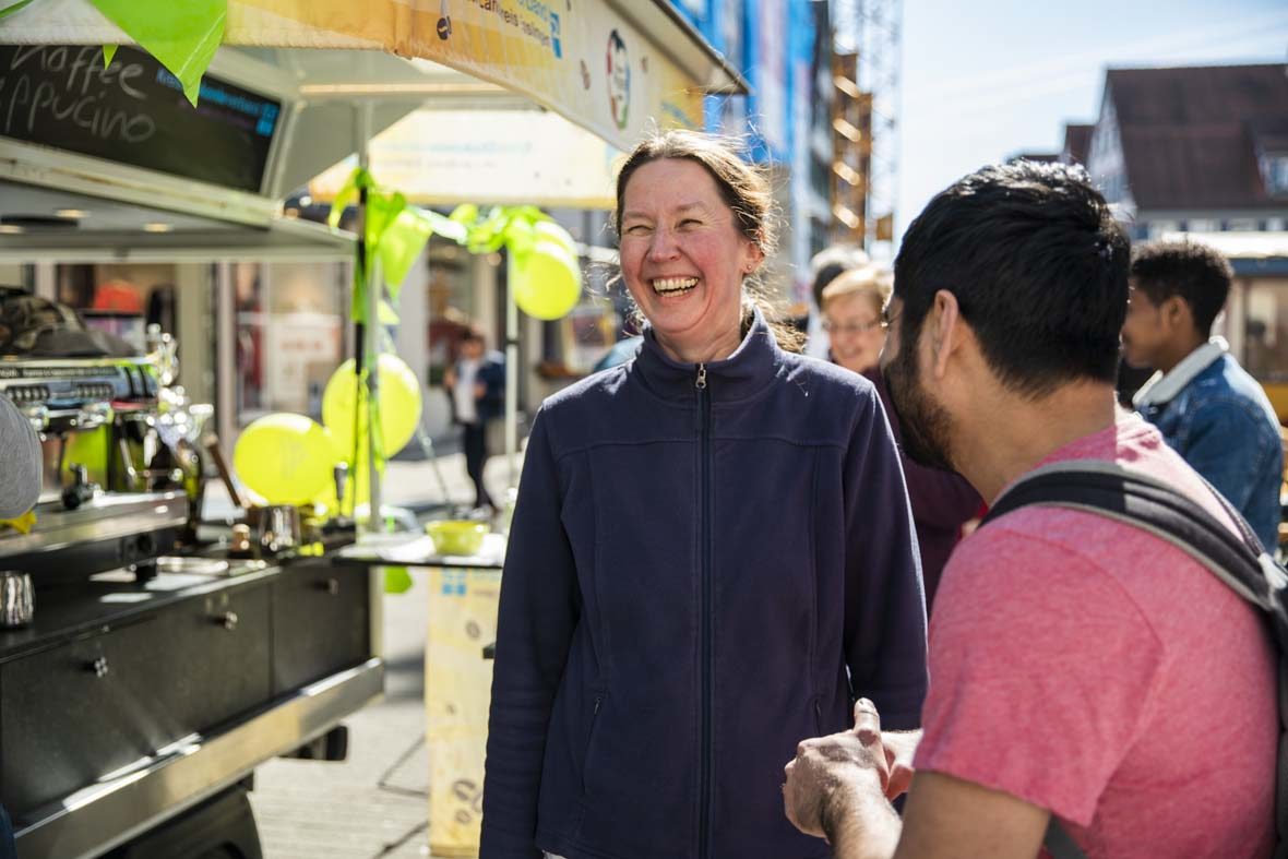 Eine Frau mit Zopf lacht herzlich. Neben ihr steht ein junger Mann in T-Shirt, mit dem sie sich unterhält. Hinter ihr sieht man das Café-Tee-Mobil, das mit Luftballons geschmückt ist.