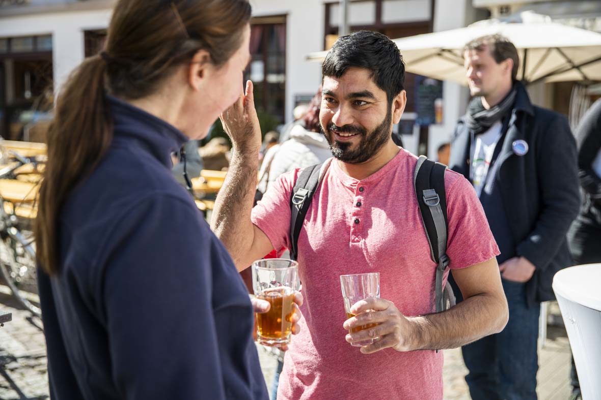 Ein junger Mann spricht mit einer Frau, er gestikuliert mit der rechten Hand und hält in der linken Hand ein Glas mit Tee. Der Mann lächelt.