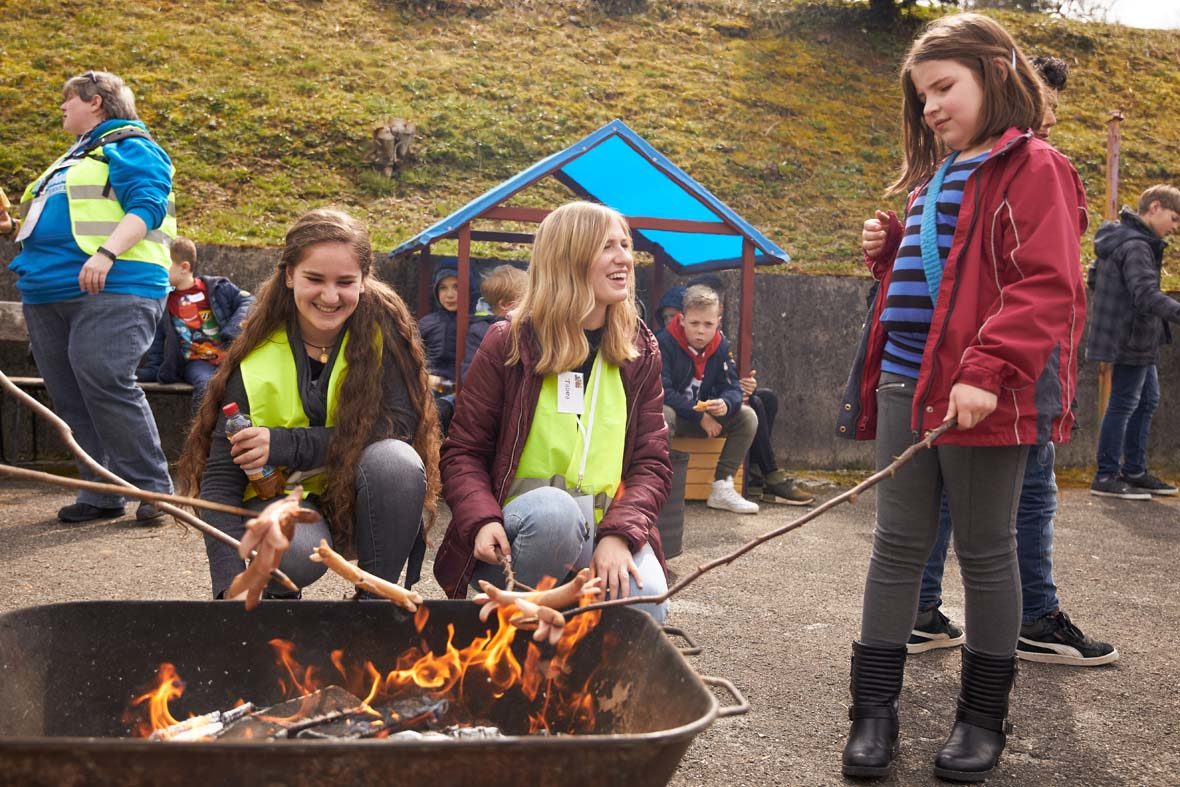 In einer eisernen Wanne ist glühende Kohle und Feuer, ein Mädchen steht rechts daneben und hält ein Würstchen am Stock darüber. Zwei Frauen in Warnweste knien vor der Wanne und eine schaut das Mädchen lachend an.