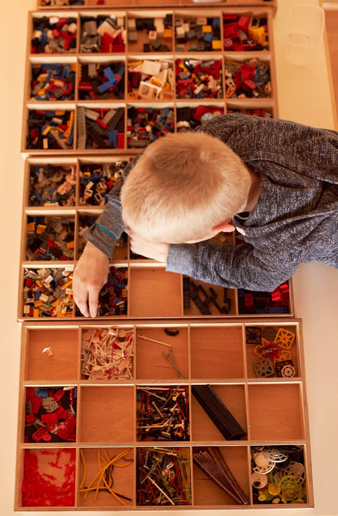 Blick von oben: Ein blonder junge lehnt über Holzkästen, in denen verschiedene LEGO-Steine sortiert gelagert sind. Er greift in eines der Kästchen.
