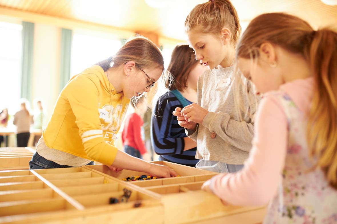 Drei Mädchen schauen in den Holzboxen mit LEGO-Steinen nach passendem Baumaterial.