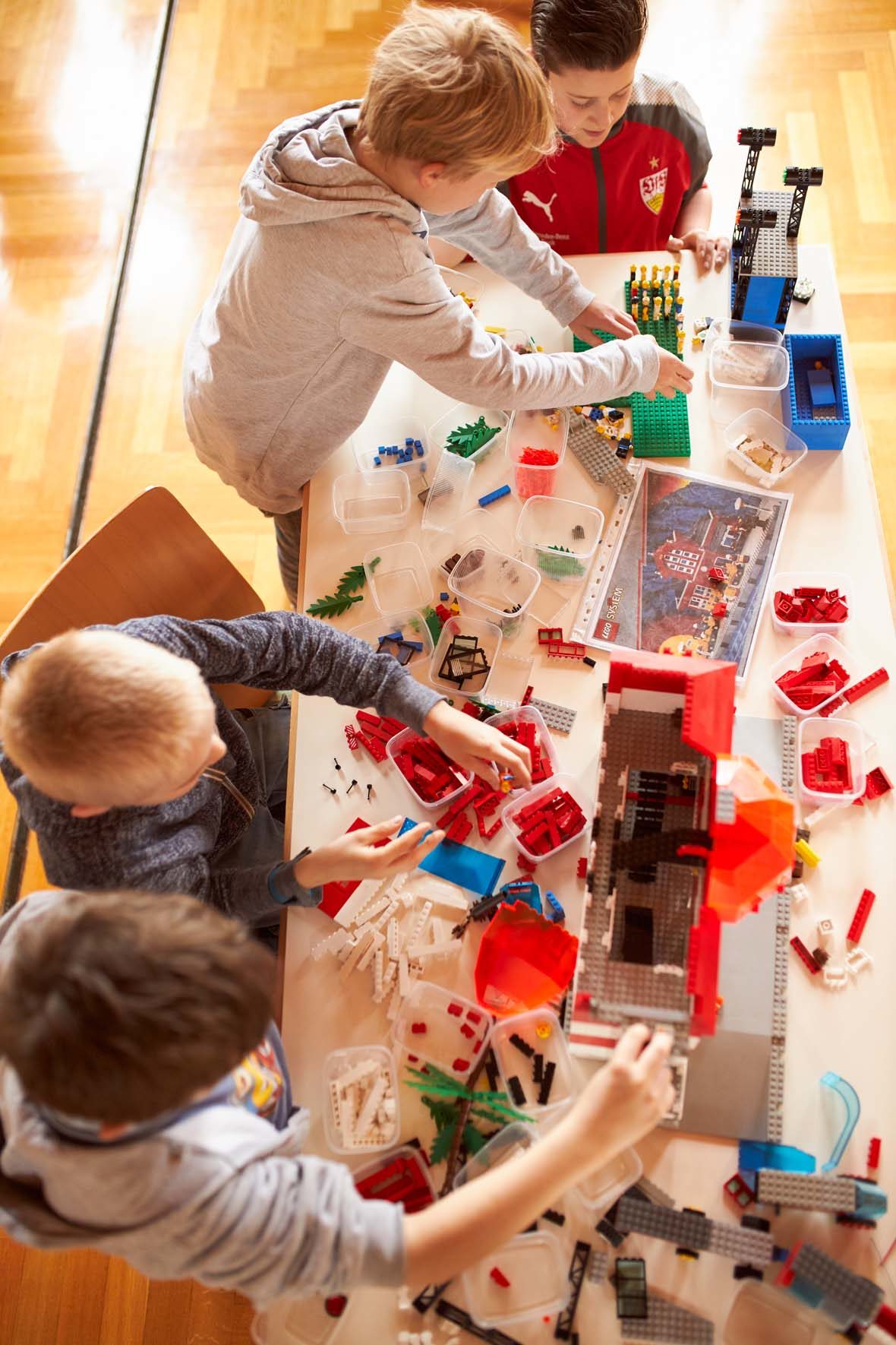 Blick von oben auf einen Tisch, dort liegen sehr viele LEGO-Steine verstreut und auch in Plastikbehältern herum. Vier Jungen bauen mit den Steinen zwei Gebäude.