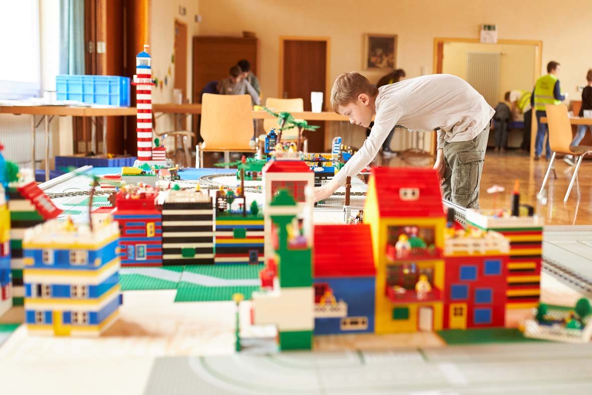 Eine LEGO-Stadt ist auf dem Bild zu sehen, im Hintergrund ein Junge, der sich darüber beugt und noch etwas baut. Weiter hinten sieht man Tische, Stühle und andere Personen.