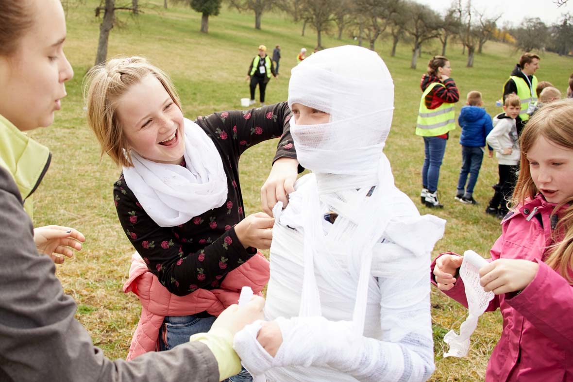 Zwei Mädchen und eine Helferin wickeln ein anderes Mädchen mit Verband ein, damit es aussieht wie eine Mumie.