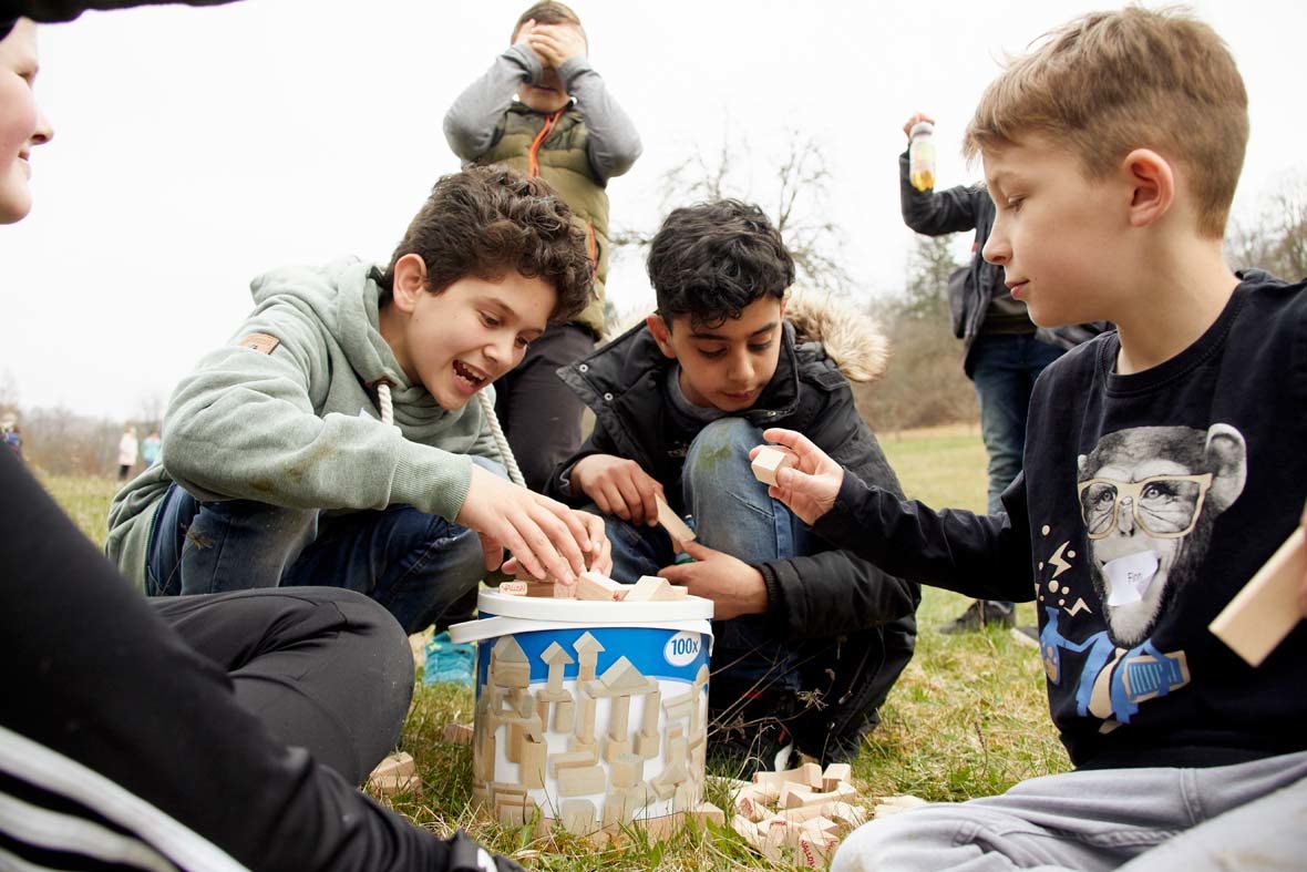 Drei Jungs sitzen vor einem Eimer mit Holzklötzen, die sie zum Teil auf der Wiese ausgebreitet haben. Im Hintergrund stehen zwei weitere Jungs, die aber nicht gut zu erkennen sind.