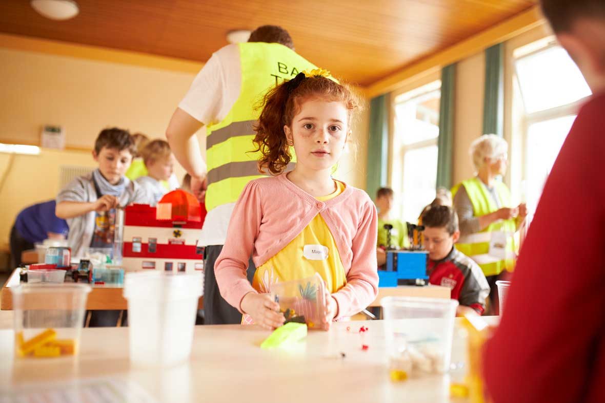 Ein Mädchen steht am Tisch und hält eine Plastikbox mit LEGO-STeinen in der Hand. Hinter ihr stehen Menschen, die an LEGO-GEbäuden bauen.