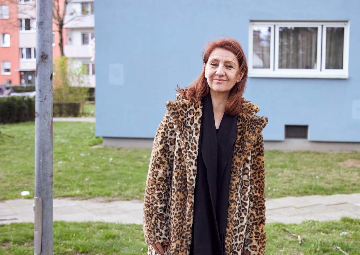 Frau mit Kunstfellmantel steht vor einem Wohnhaus.