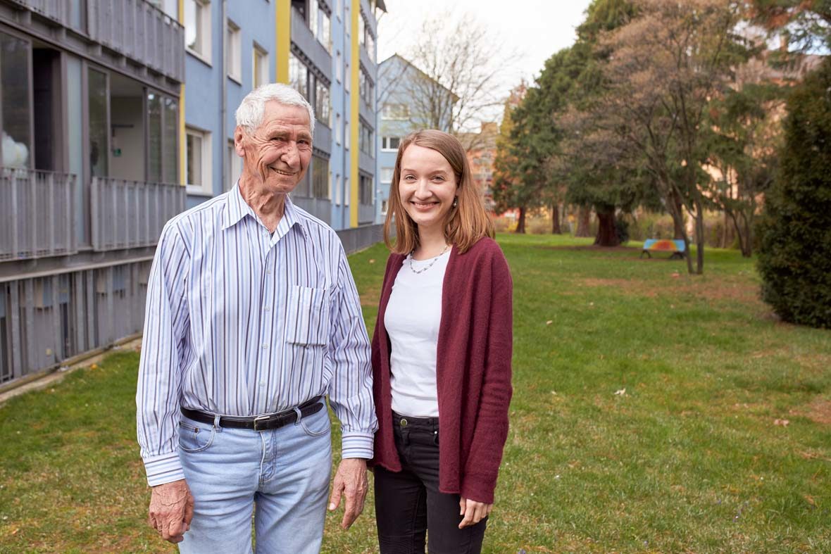 Ein älterer Mann und eine junge Frau stehen auf der Wiese vor einem Wohnhaus.