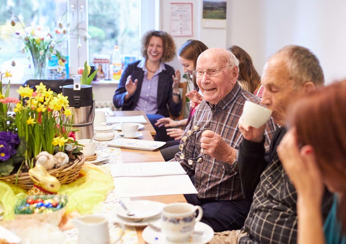 Mehrer Menschen sitzen an einem Tisch, trinken Kaffee und singen.