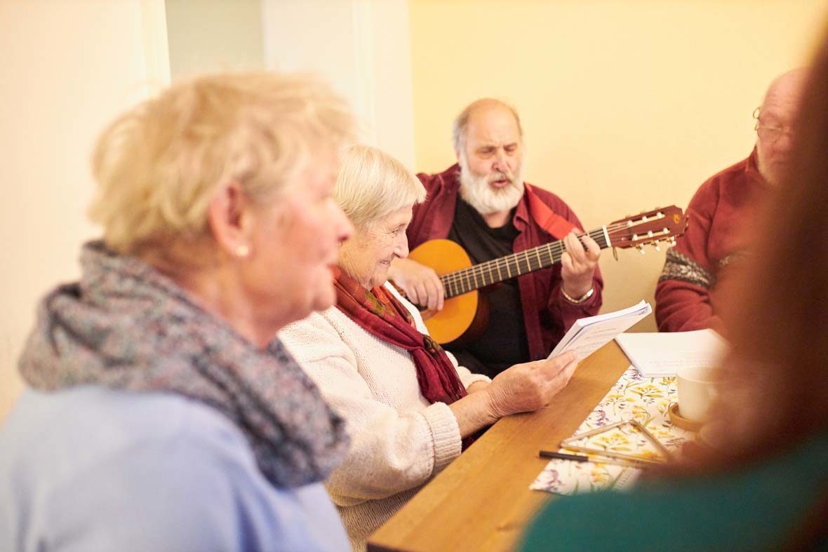 Ein älterer Mann sitzt mit mehreren Menschen am Tisch und spielt Gitarre.