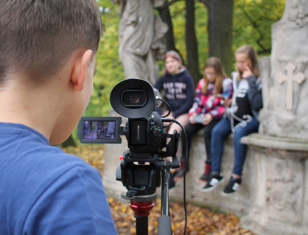 Foto: Junge steht vor Filmkamera, schaut hinein und Filmt drei Mädchen, die auf einer Mauer sitzen.