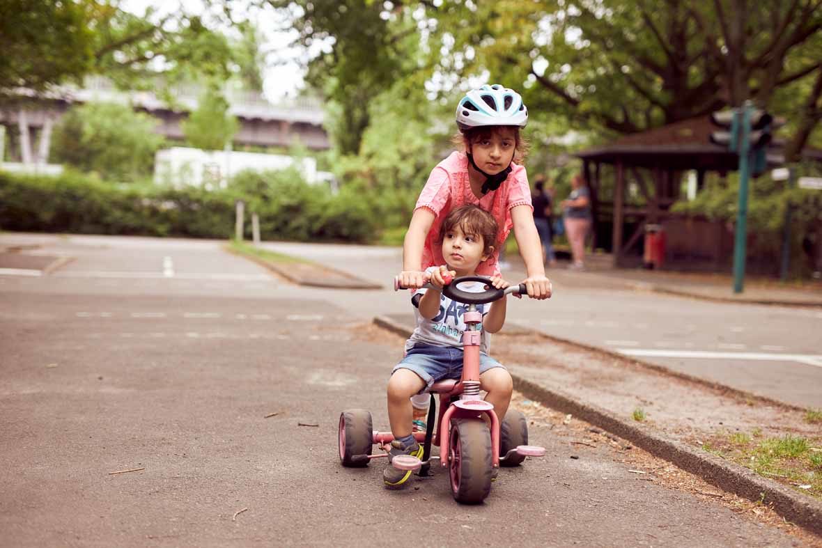 Ein Mädchen mit Helm steht über ein Dreirad gebeugt, auf dem Dreirad sitzt ein kleiner Junge