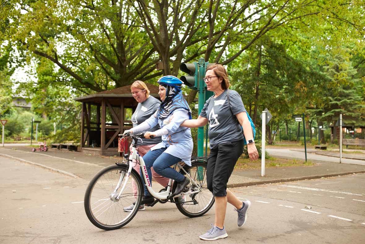 Foto: Zwei Frauen laufen neben einem Fahrrad her und stützen die Fahrradfahrerin
